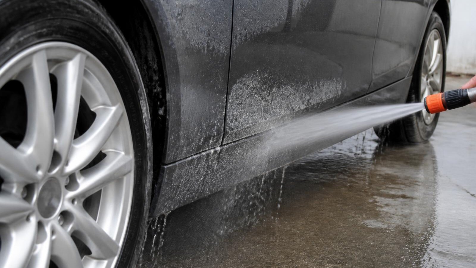 A close-up of an alloy wheel and lower rocker panel being rinsed with a hose, salt film washing away.