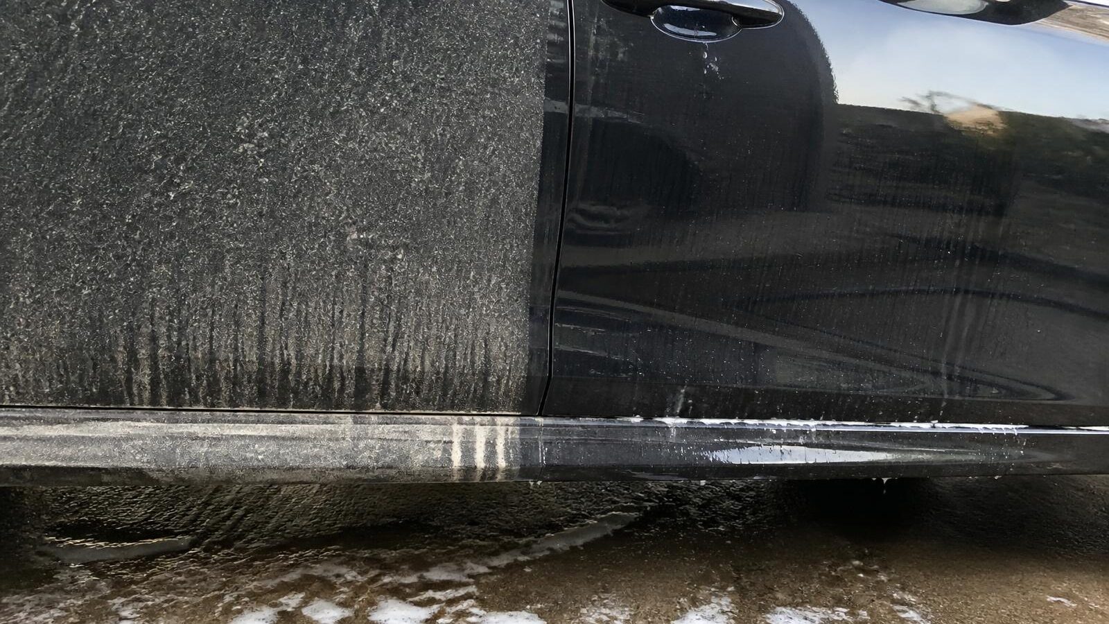 A car door and rocker panel photographed mid-wash: one half still covered in grey road film, the other half washed and glossy.